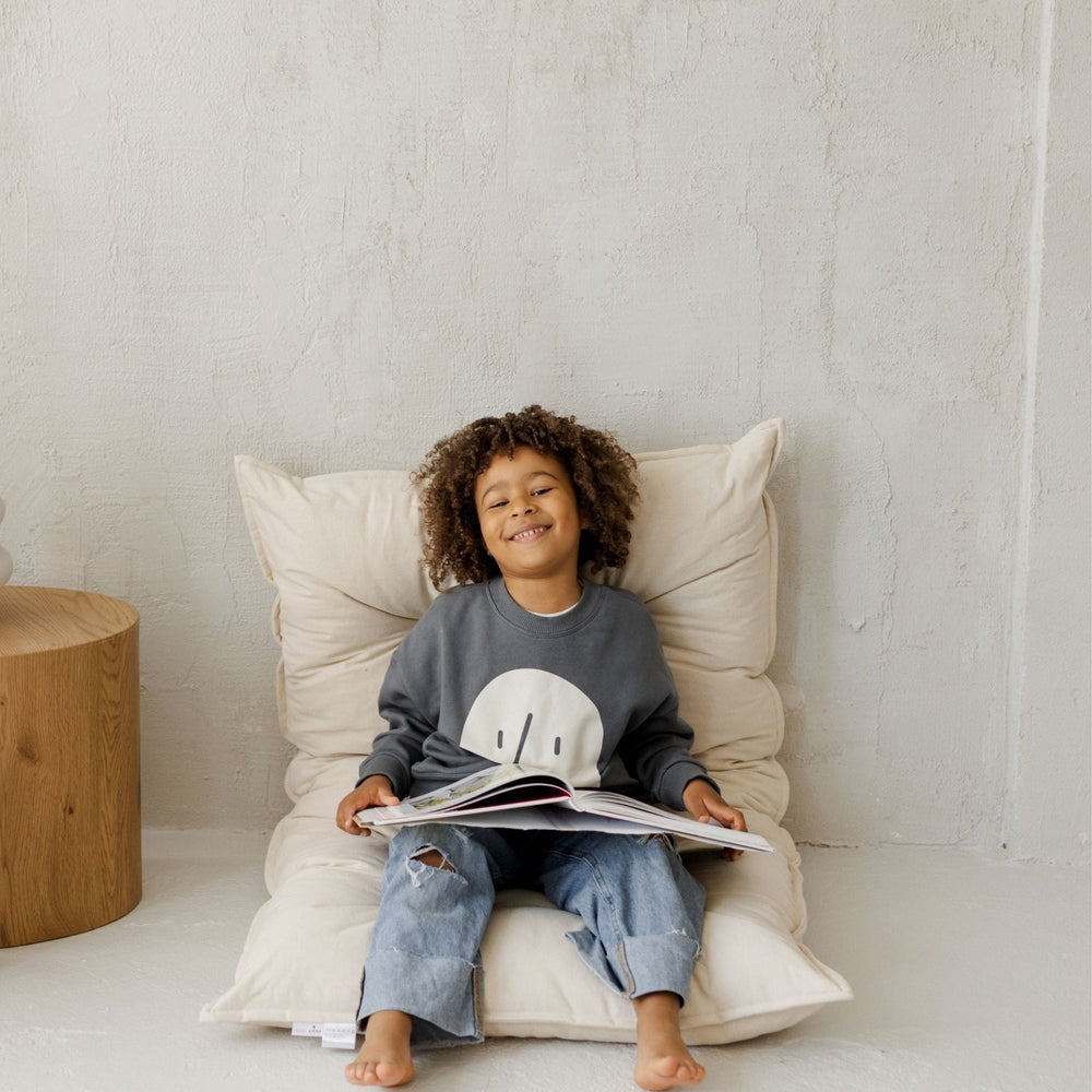 Child sitting on a large pillow reading a book in a cosy room.