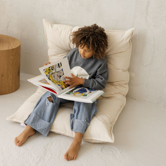 Child sitting on a folded floor cushion so it's like a chair reading a book in a cosy room.