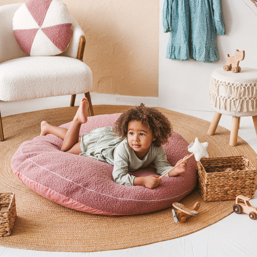 Child lying on a pink floor cushion in a cosy room with toys and furniture.