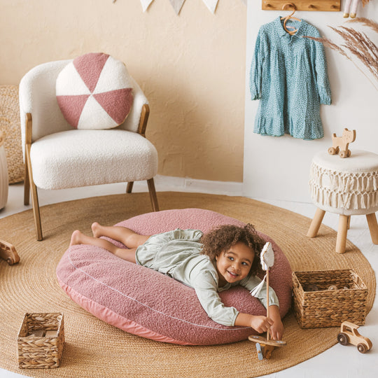 Child lying on a pink cushion in a cosy room with a chair, toys, and a dress on a rack.