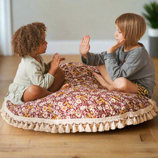 Two children sitting on a floral round cushion with tassels playing a clapping game