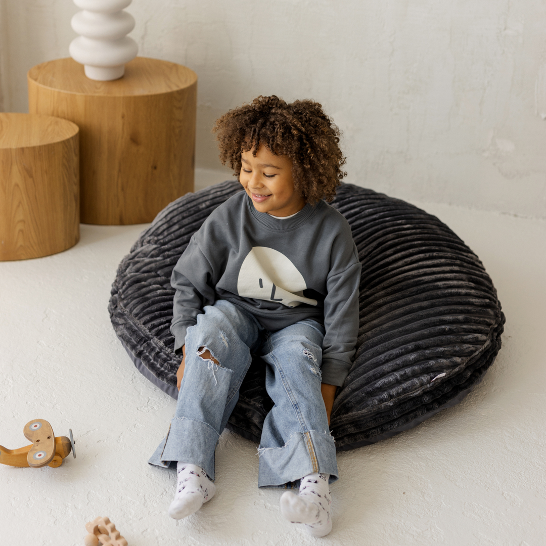 Child sitting on a textured floor cushion in a minimalistic room with wooden tables and a white wall.