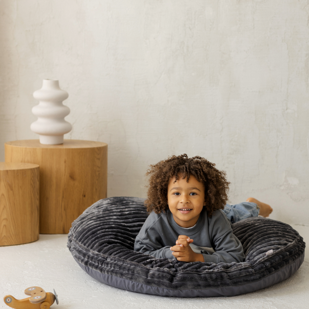 Child lying on a grey round floor cushion in a room with wooden stools and a white wall.