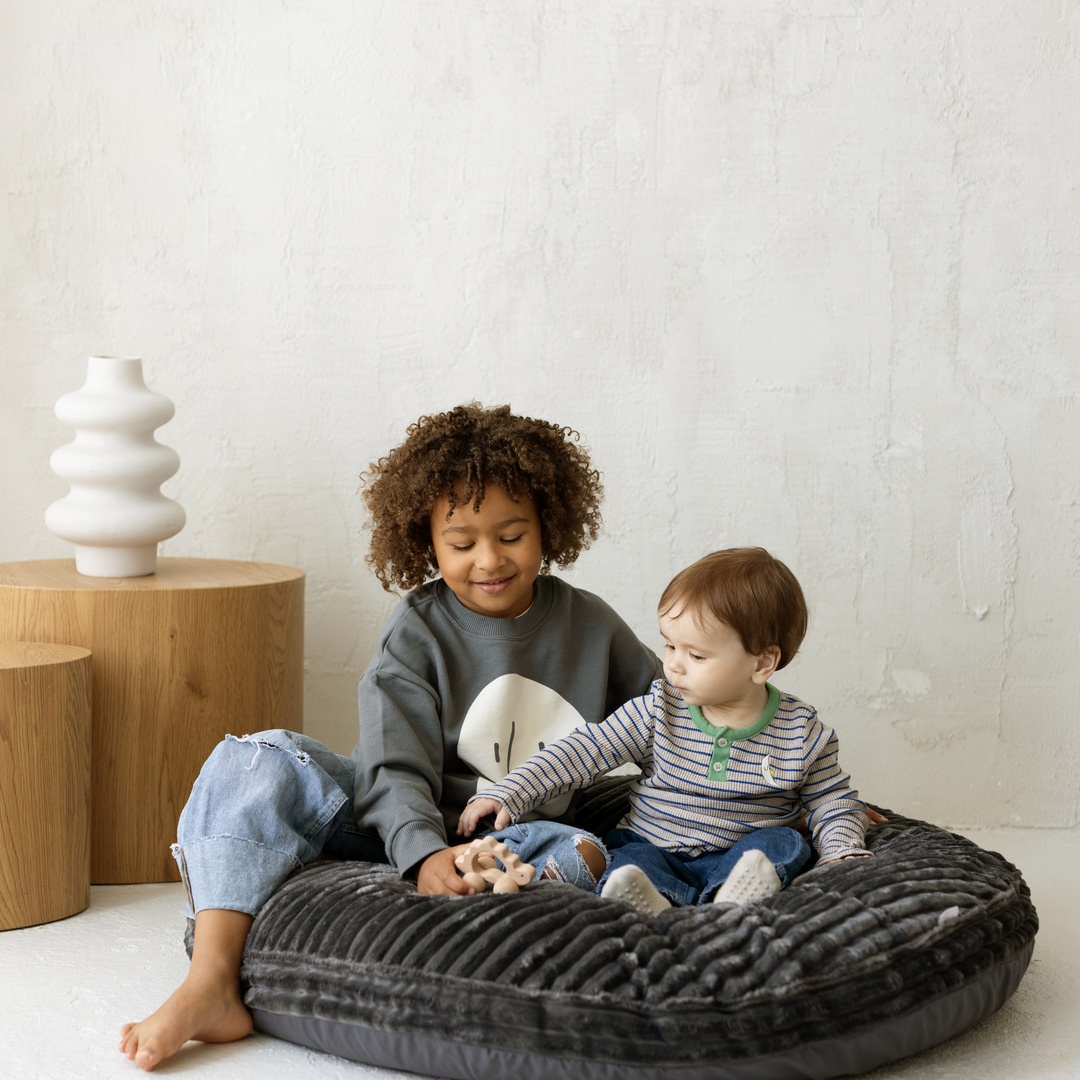 Two children sitting on a large grey floor cushion in a room with wooden tables and a white wall.