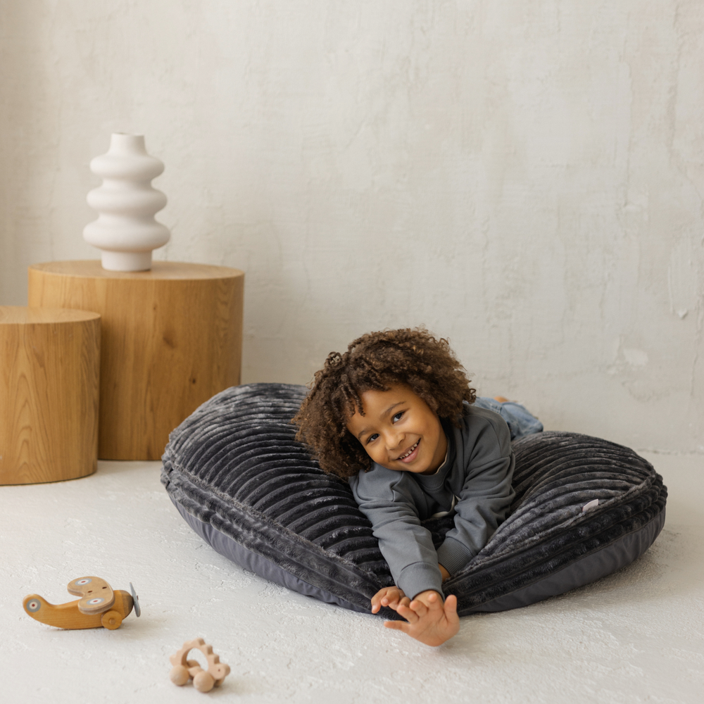 Child lying on a large grey cushion with wooden toys and a white vase in the background