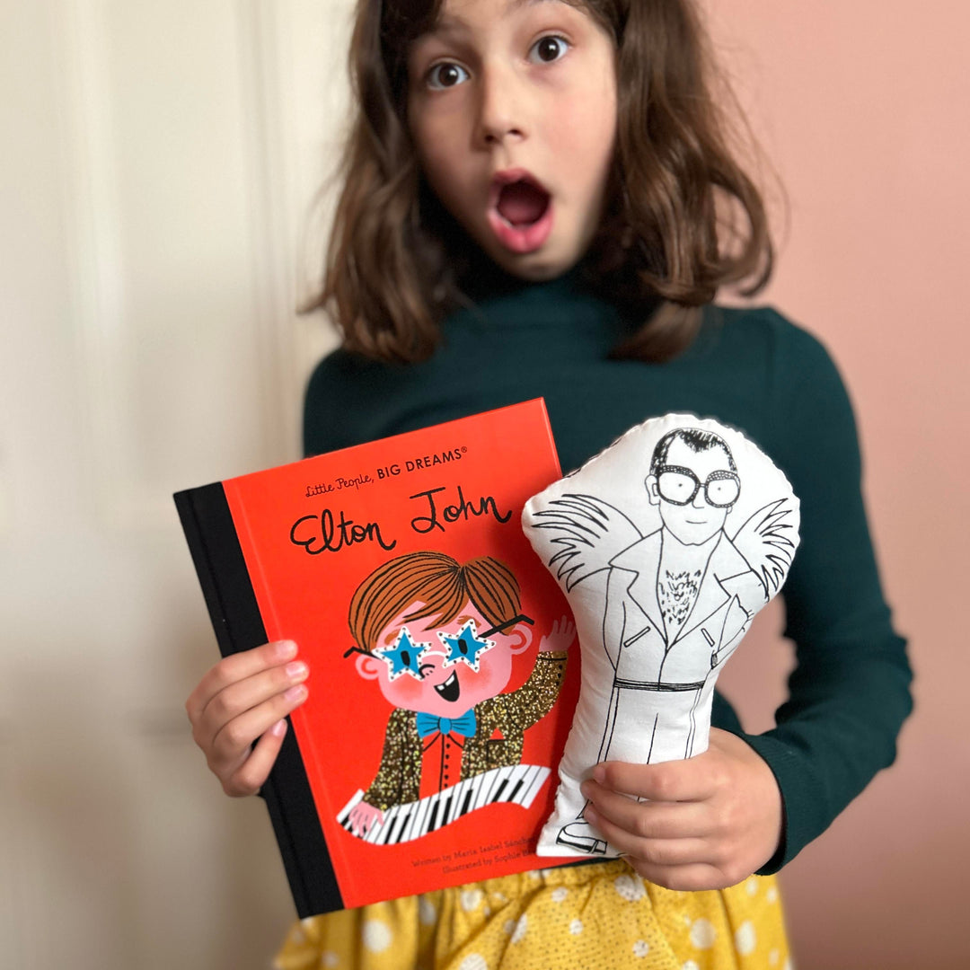 A child holding a screen printed doll of Elton John and a red book of Elton John with a surprised look on her face