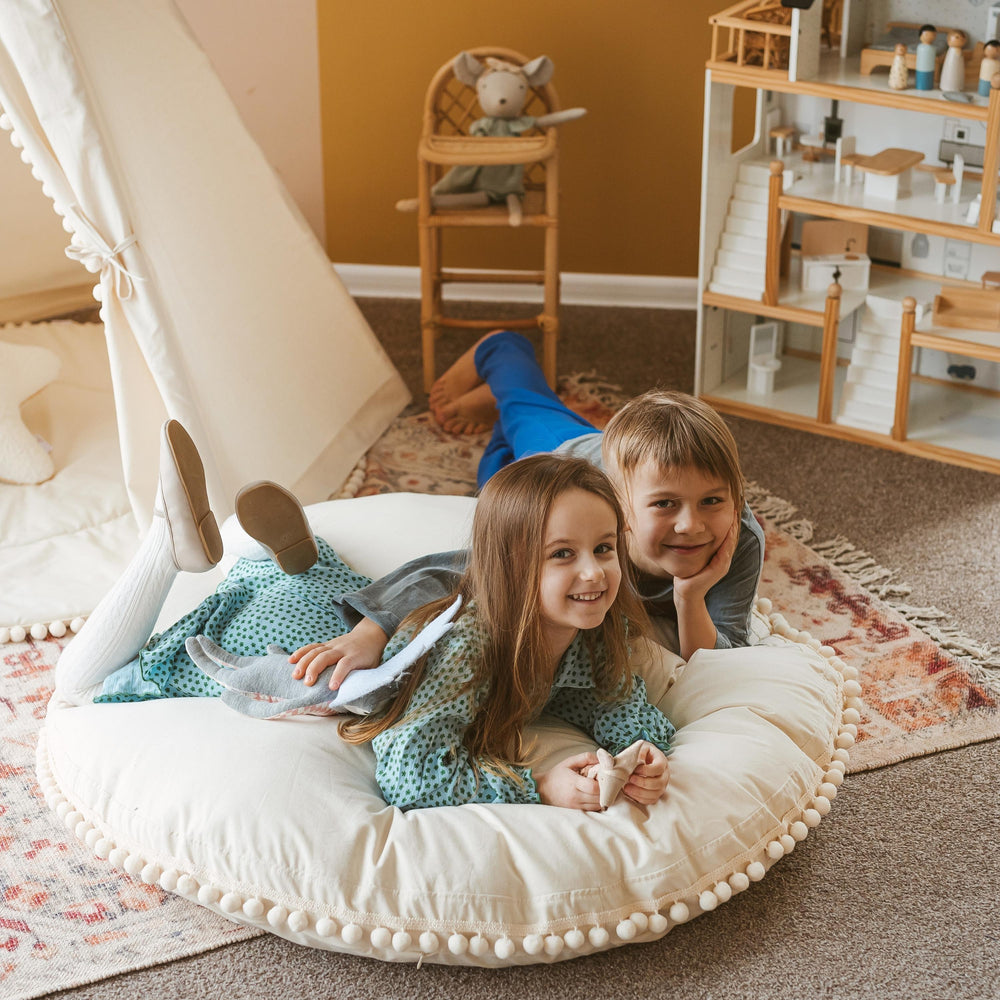 Two children lying on a round white cushion in a playroom with toys and furniture.
