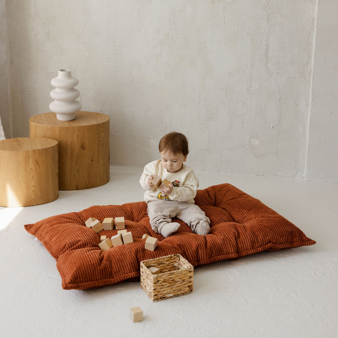 Child playing on a large brown floor cushion with wooden blocks and a woven basket in a minimalistic room.