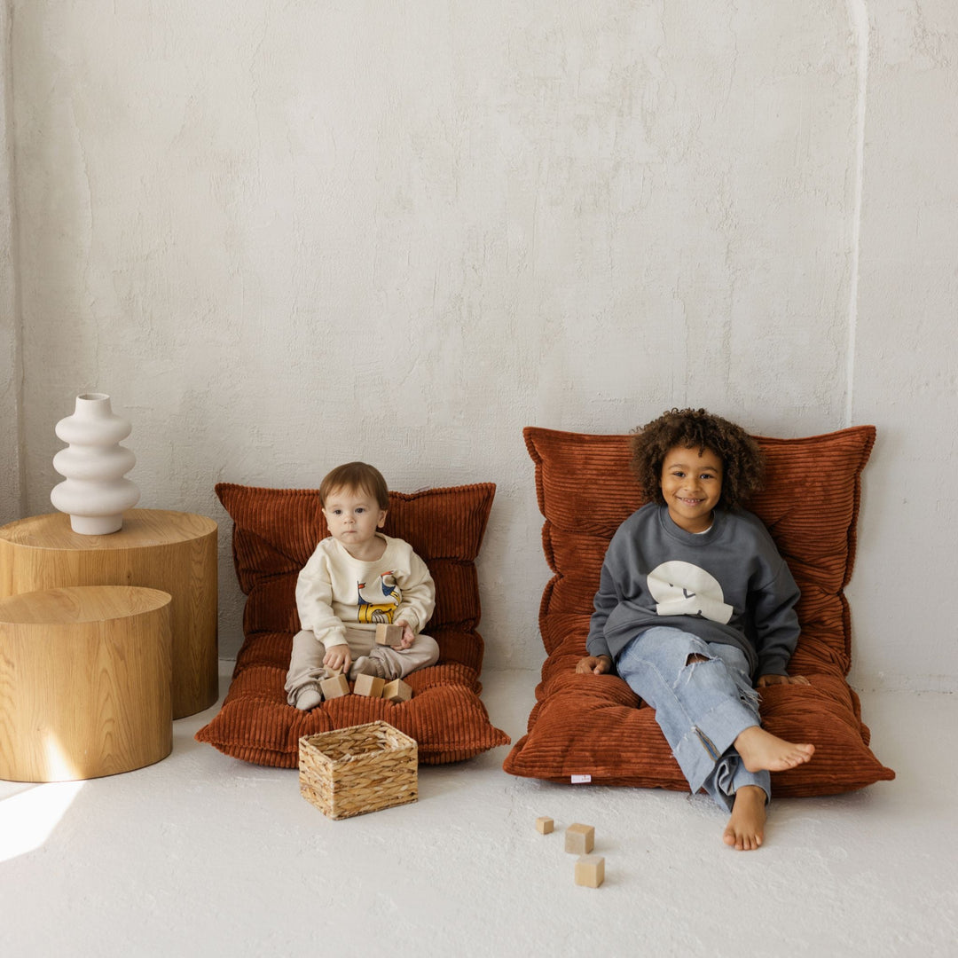 Two children sitting on large brown cushions in a room with wooden tables and toys.