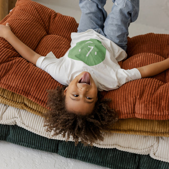 Child lying on a stack of colourful cushions with a white shirt and blue jeans.