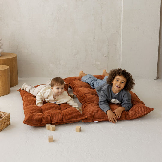 Two children playing on a large brown cushion in a minimalistic room.