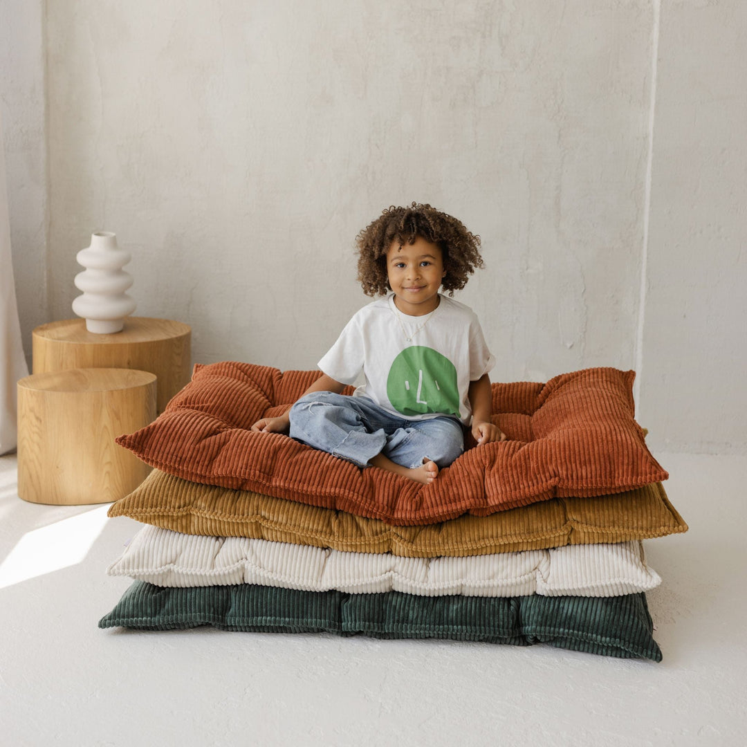 Child sitting on a stack of colourful cushions in a minimalistic room.