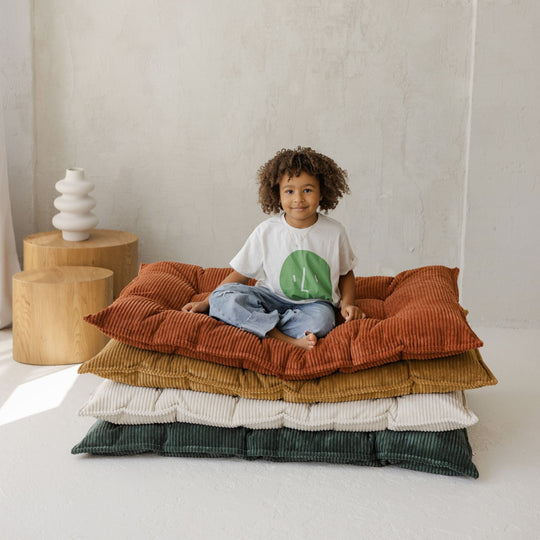 Child sitting on a stack of colourful cushions in a minimalistic room.