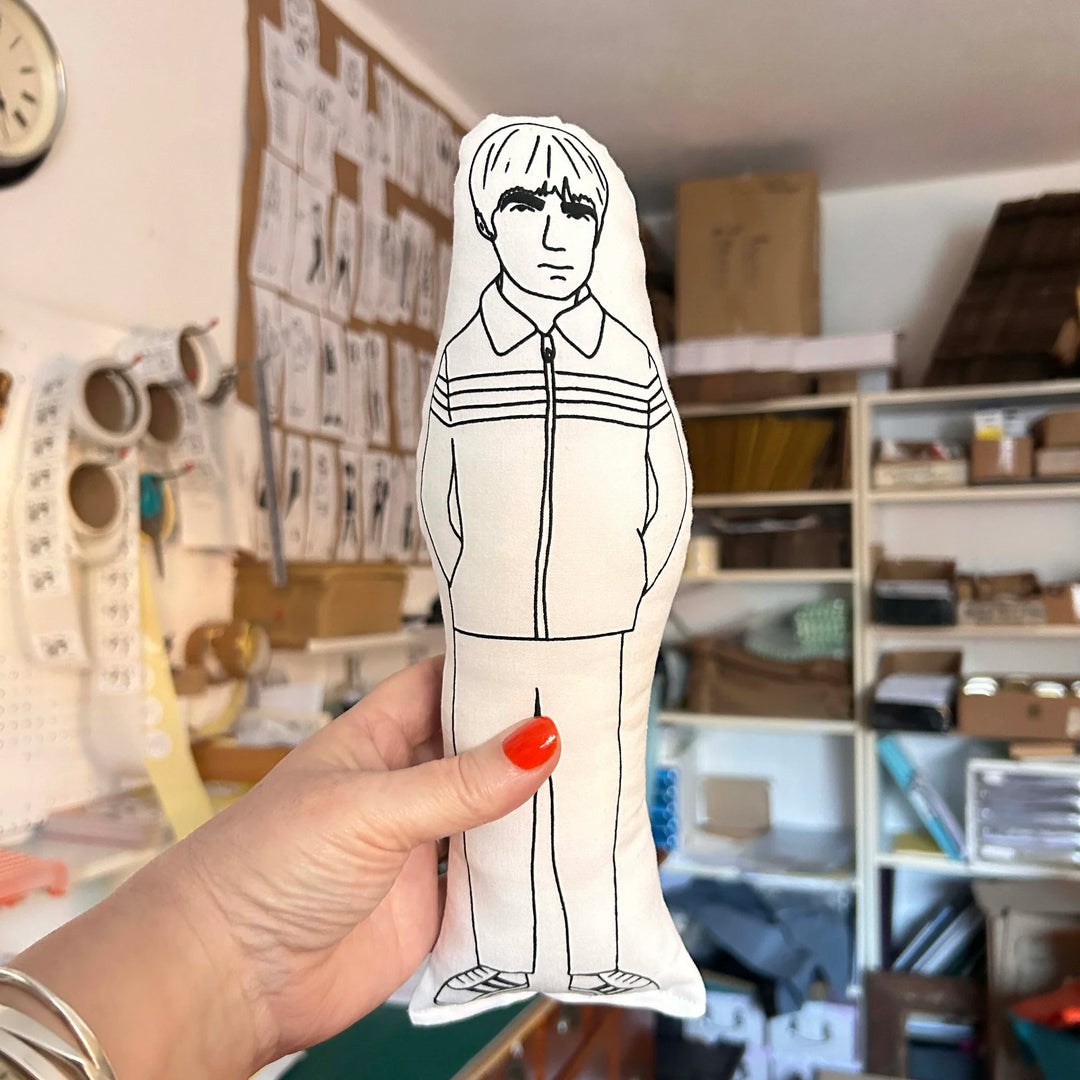 Hand holding screen printed doll cushion of Noel Gallagher against backdrop of out of focus interior room