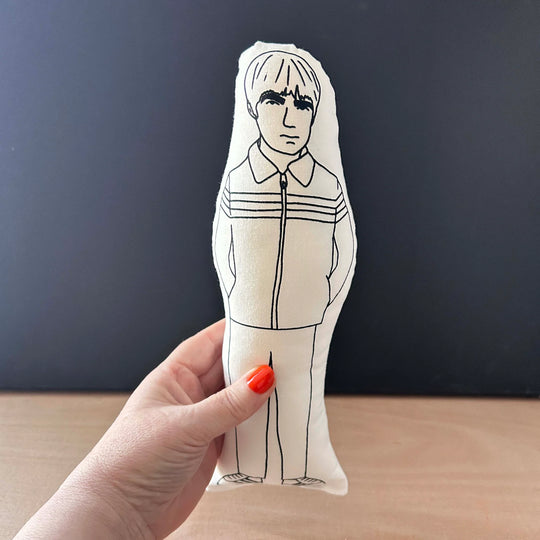 Hand holding a screen printed doll cushion with white fabric and black illustration depicting Noel Gallagher wearing a zip-up shell suit jacket
