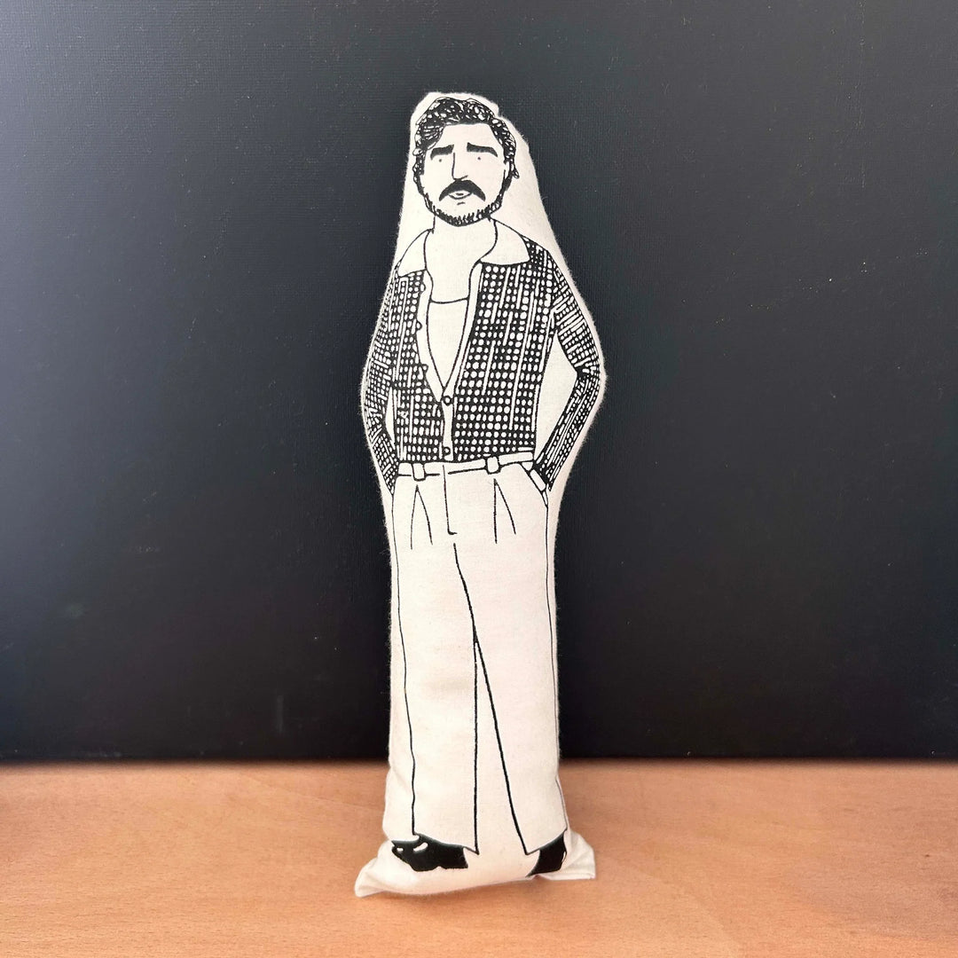 Screen printed doll cushion with white fabric and black illustration depicting Pedro Pascal wearing smart trousers, black shoes and an open checked shirt with his hands in his pockets