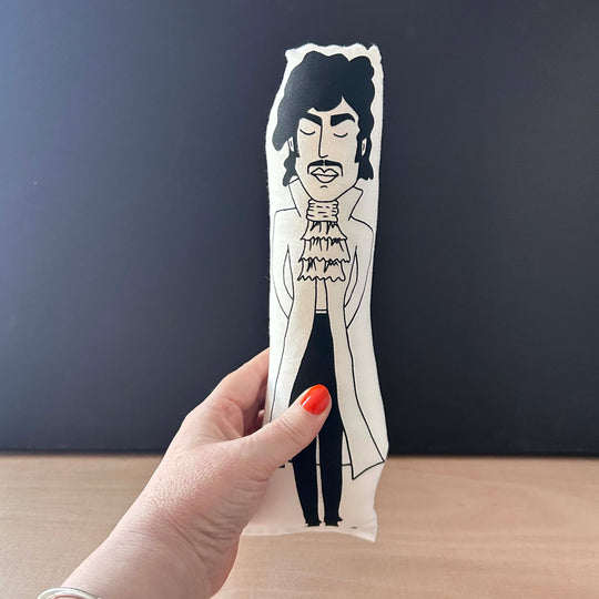 Hand holding a screen printed doll cushion with white fabric and black illustration depicting Prince  wearing a long high collared formal coat with a frilled crevate with his hands behind his back and his eyes closed