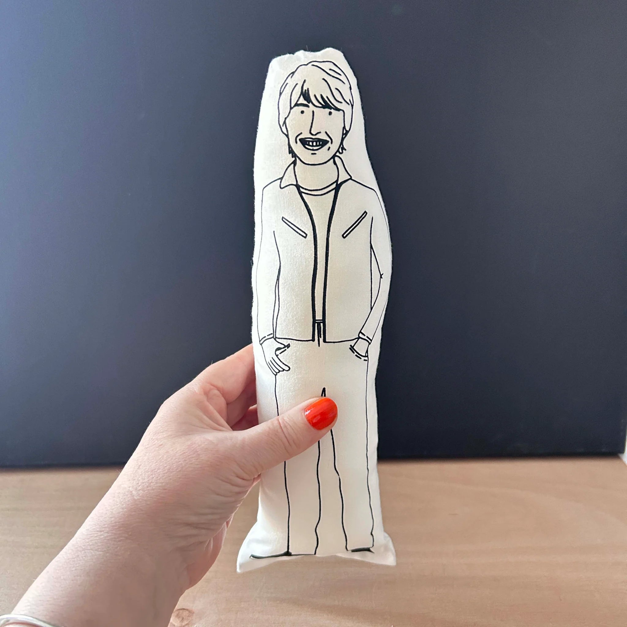 Hand holding screen printed doll cushion with white fabric and black illustration depicting physicist and TV presenter Professor Brian Cox, smiling wearing a casual outfit with his hands in his pockets