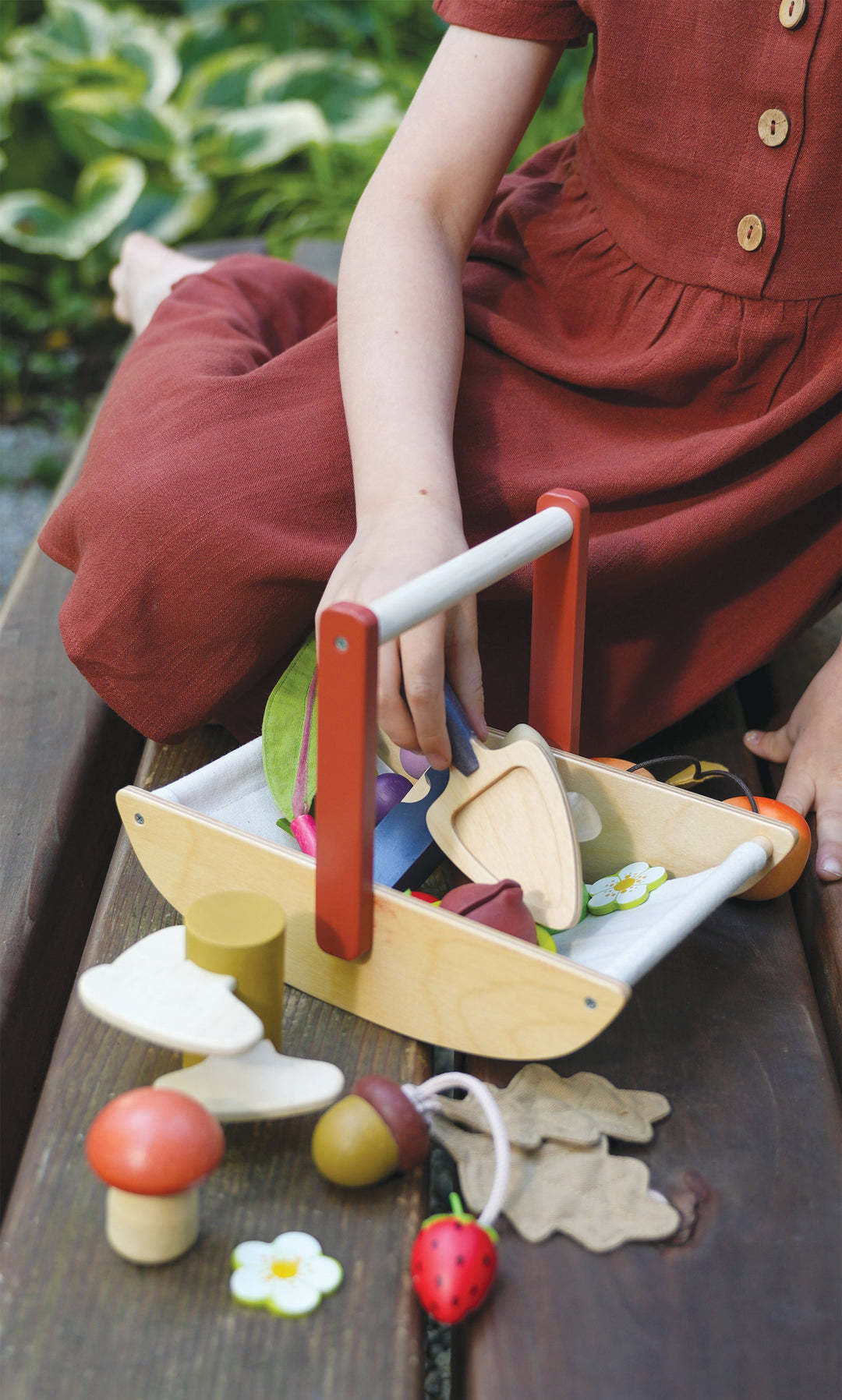 Child playing with a wooden toy gardening trug filled with colourful wooden fruits and vegetables on a wooden surface.