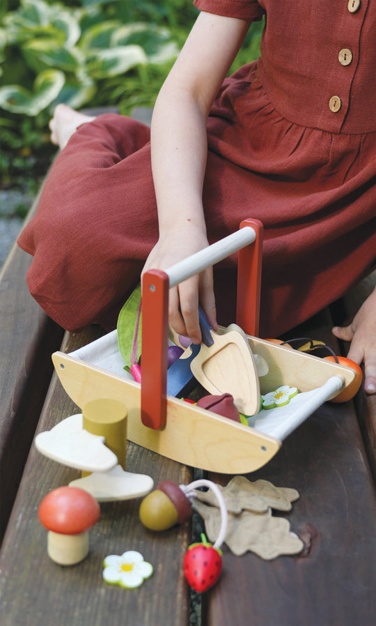 Child playing with a wooden toy gardening trug filled with colourful wooden fruits and vegetables on a wooden surface.