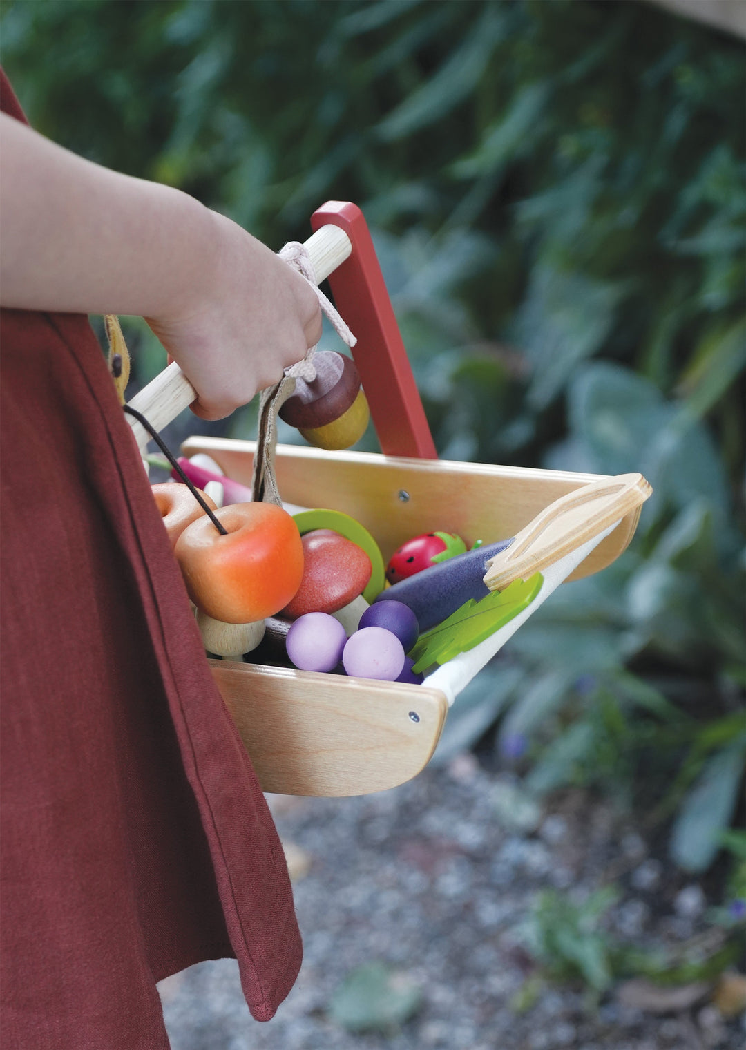 Child holding a wooden basket with toy fruits and vegetables outdoors.