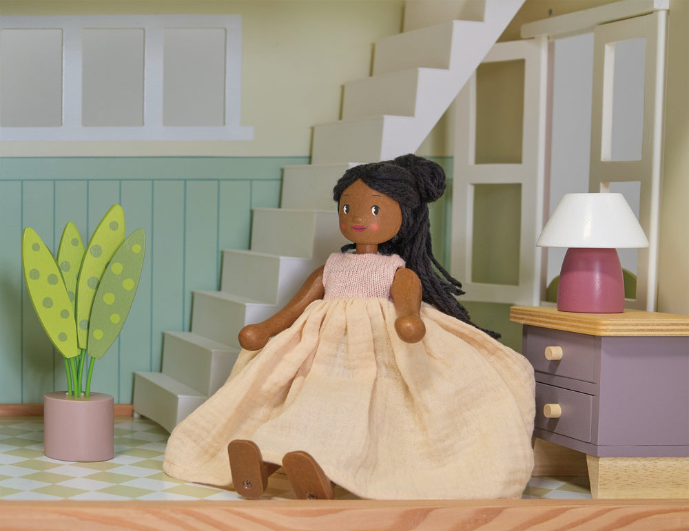 Doll in a beige dress sitting on a wooden floor with a staircase and lamp in the background.
