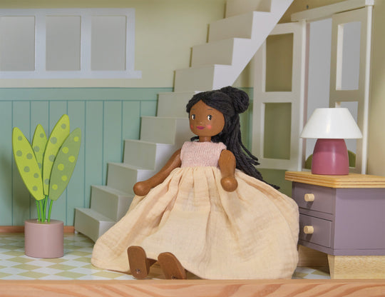 Doll in a beige dress sitting on a wooden floor with a staircase and lamp in the background.
