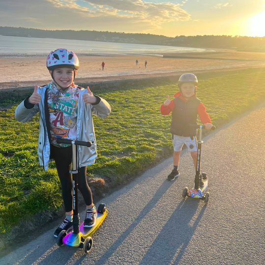 Two children on scooters by a road with a scenic background