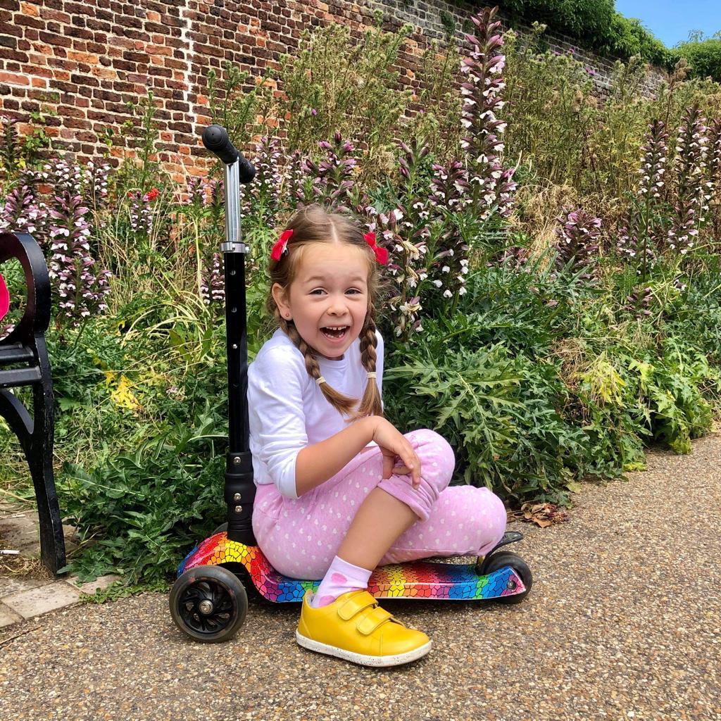 Young girl sitting on a scooter in a garden setting with flowers and a brick wall in the background.