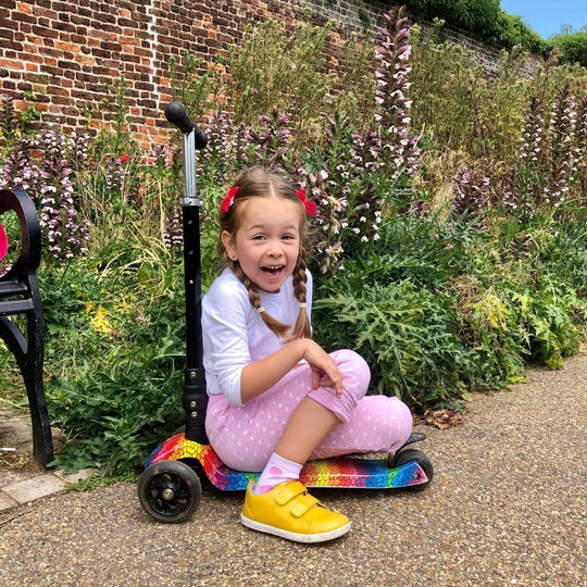 Young girl sitting on a scooter in a garden setting with flowers and a brick wall in the background.