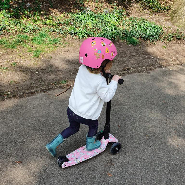Child wearing a pink helmet and white jacket riding a scooter on a paved path.