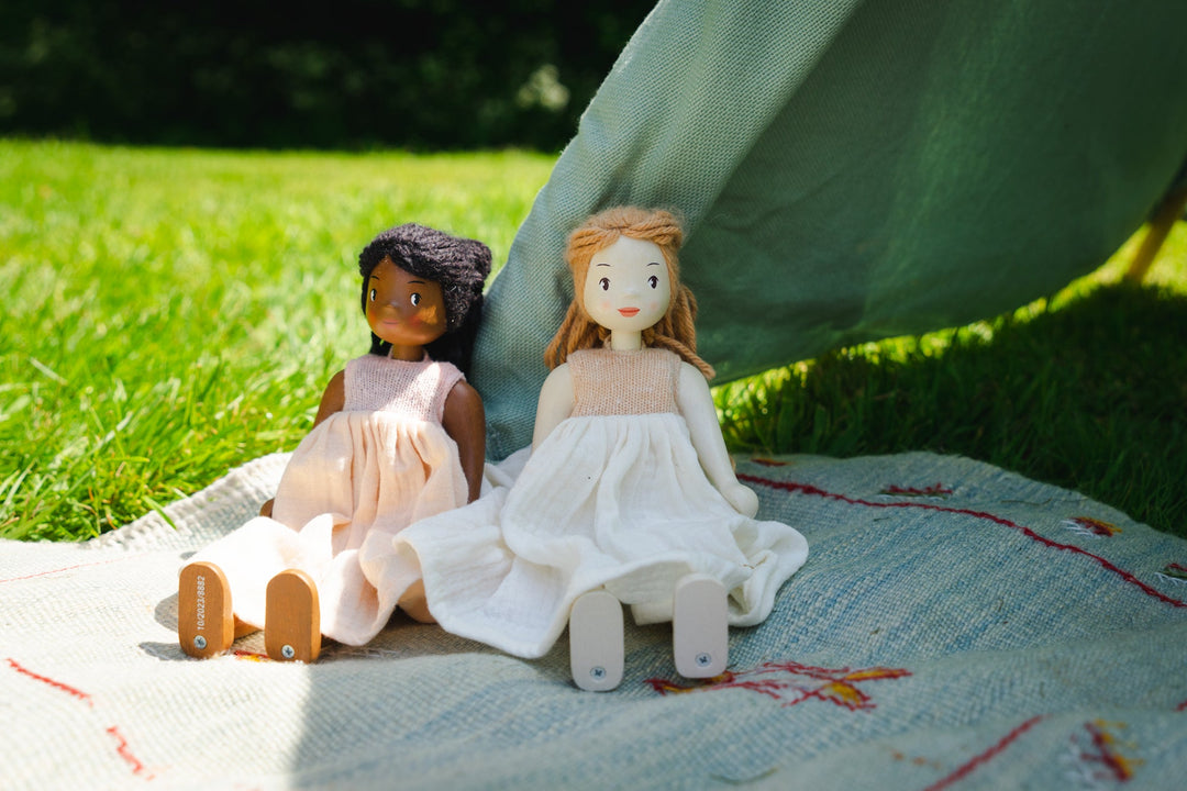 Two dolls in dresses sitting on a blanket outdoors.