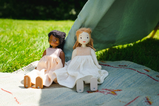 Two dolls in dresses sitting on a blanket outdoors.