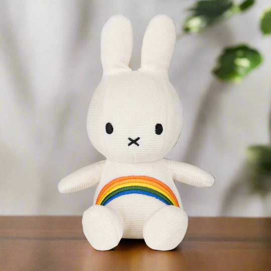 White plush toy rabbit with iconic cross stitched mouth, with white corduroy fabric and an embroidered rainbow across the chest