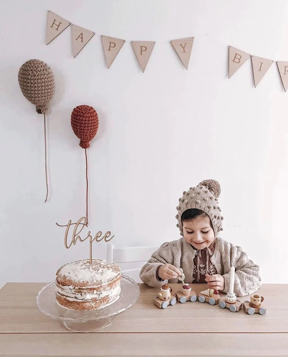 A chld sat at a wooden table playing with a wooden stacking train toy with a happy birthday banner on the wall behind with two different sized crochet balloons as wall decor