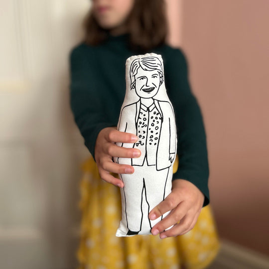 A child holding out a black and white screen printed cushion doll depicting Sandi Toksvig towards the camera with a blurred background