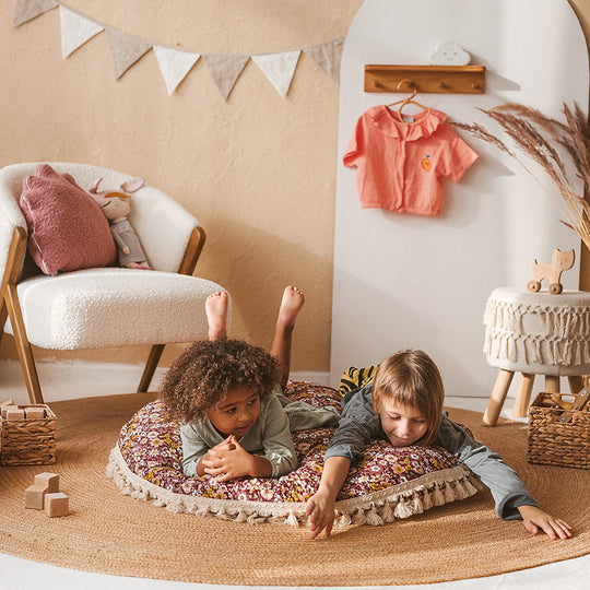 Two children playing on a round floor cushion room with a chair, clothes, and decorative elements.