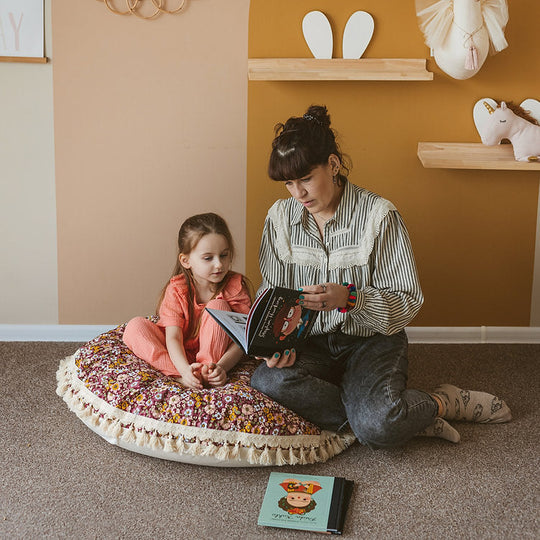 Woman and child sitting on a round floral floor cushion reading a book in a cosy room.