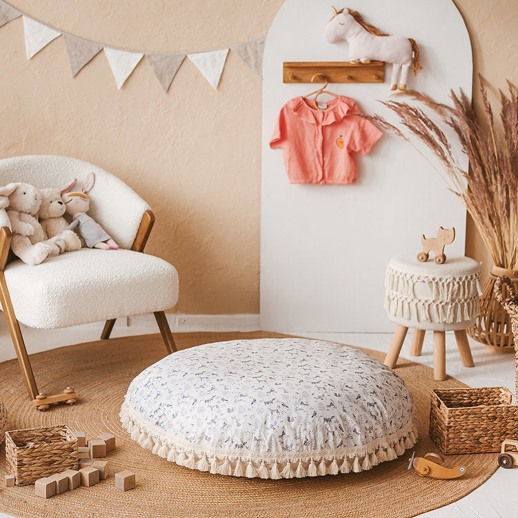 Children's room with toys, a plush chair and a large round floor cushion which is white with a pattern animal design and a white trim with tassels