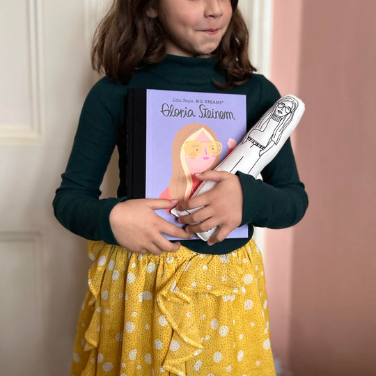 A child holding a screen printed doll cushion of Gloria Steinem and a Little People Big Dreams book about the life of Gloria Steinem