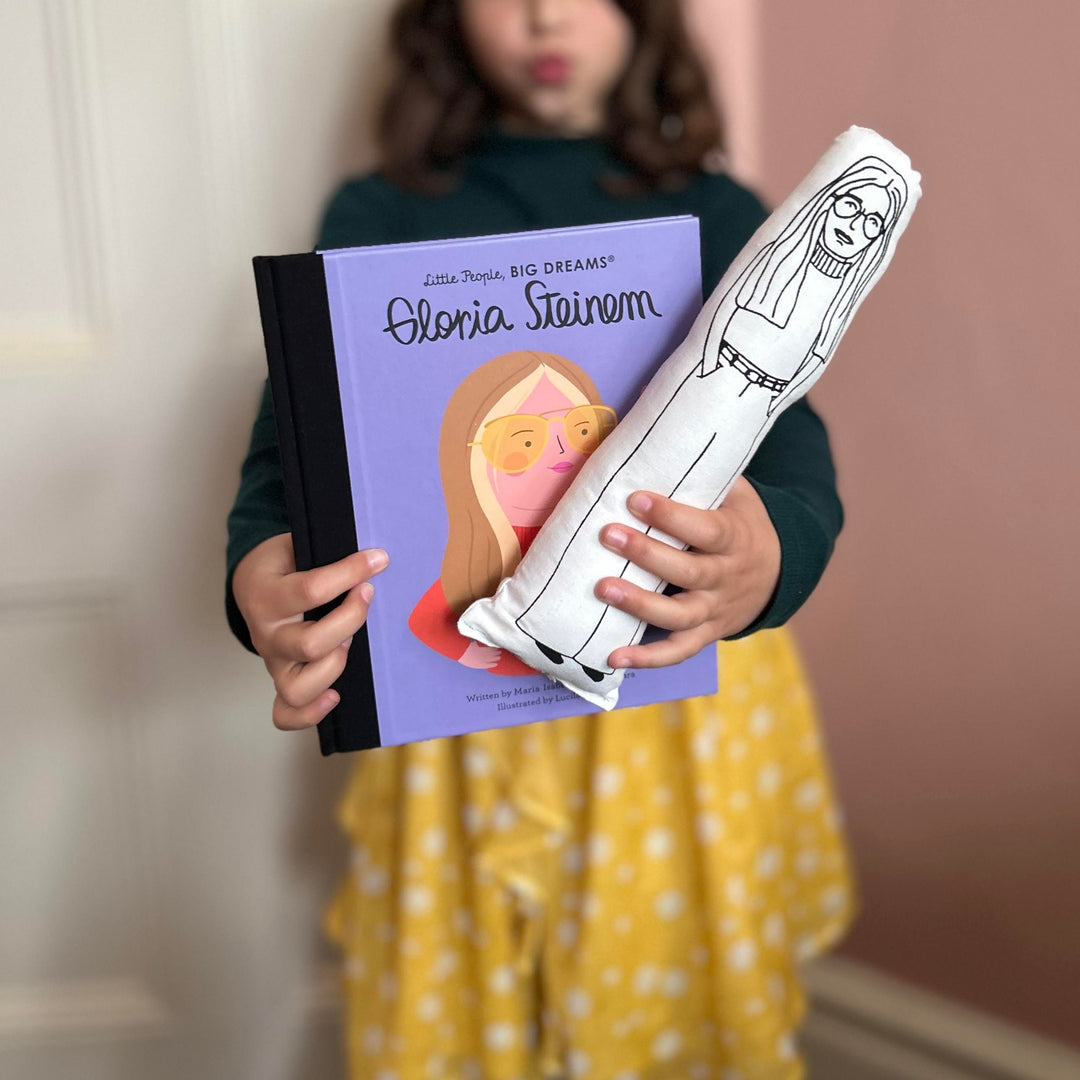 A child holding a black and white screen printed cushion doll with an illustration of Gloria Steinem, and a book with the same cover.