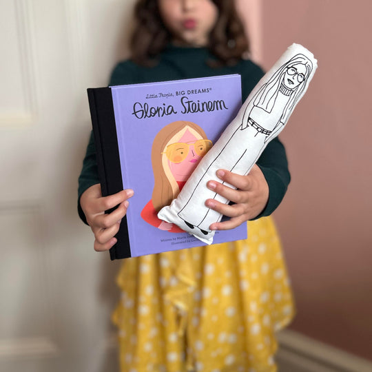 A child holding a black and white screen printed cushion doll with an illustration of Gloria Steinem, and a book with the same cover.