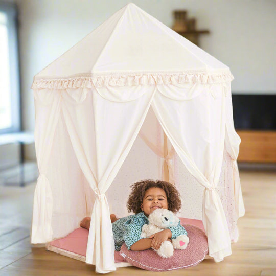 Child lying on a play mat inside a white pavilion shaped play tent cuddling a teddy bear