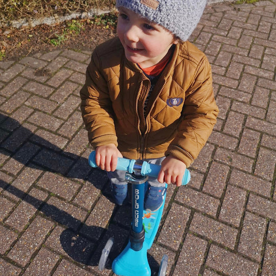 Child on a blue scooter wearing a brown jacket and grey beanie on a paved surface.