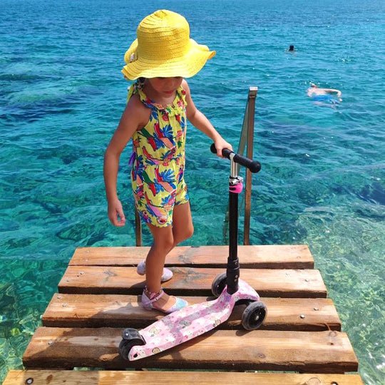 Child with a colourful swimsuit and yellow hat standing on a wooden dock by clear blue water, holding the scooter.