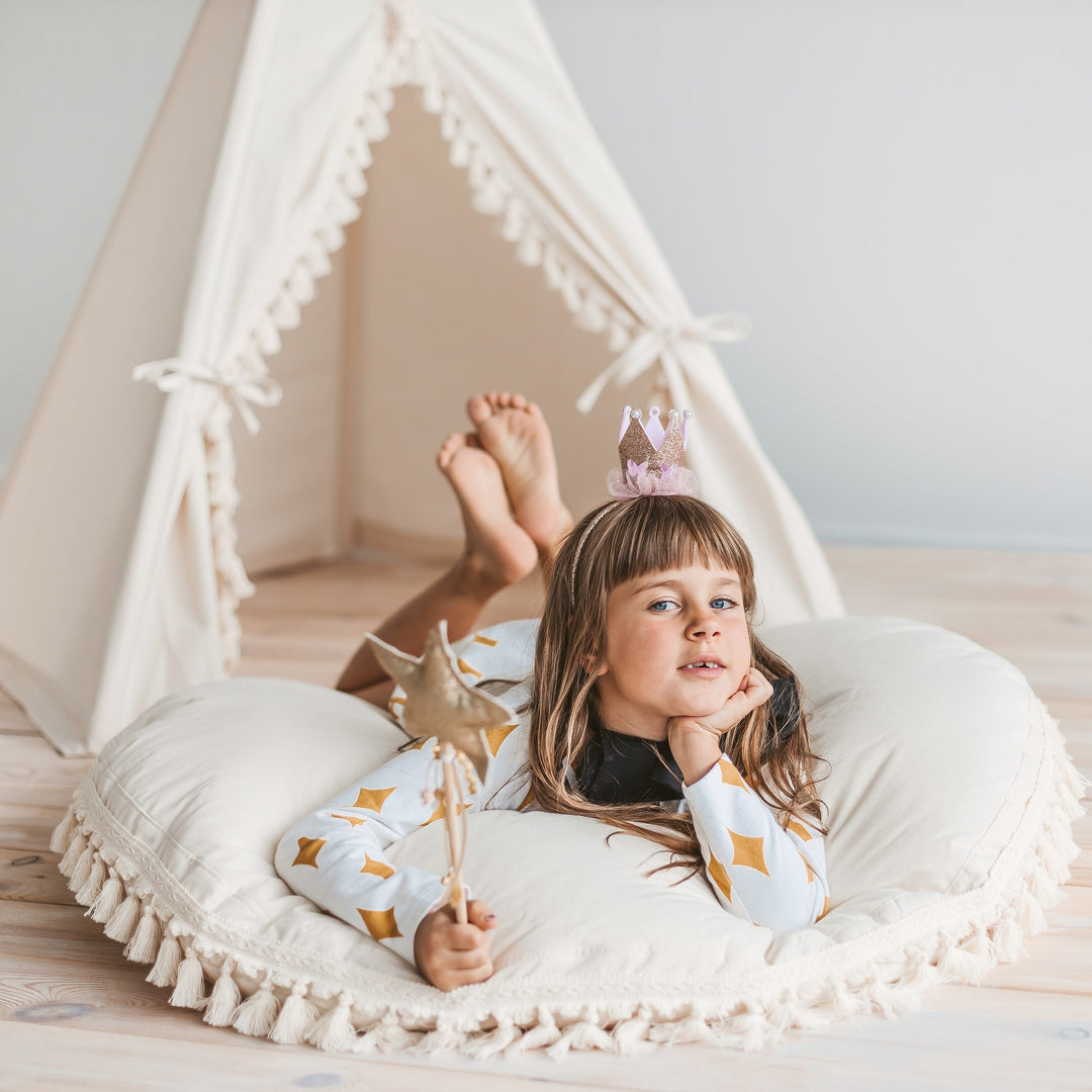 Child lying on a floor cushion in front of a teepee with a crown on, holding a wand.
