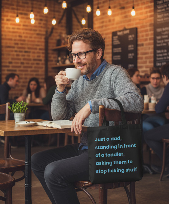 Man sitting at a table in a cafe holding a cup, with a humorous message on his tote.