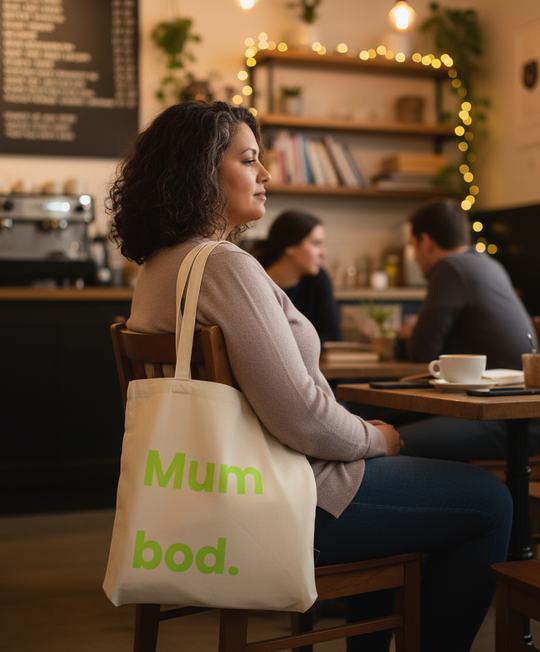 Woman sitting in a cafe with a tote bag labeled 'Mum bod.'