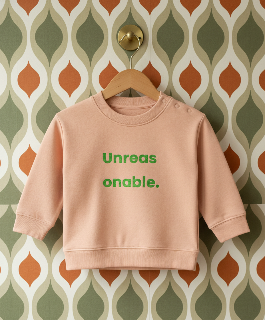 Pink sweatshirt with green text 'Unreas onable.' on a hanger against a patterned wall.