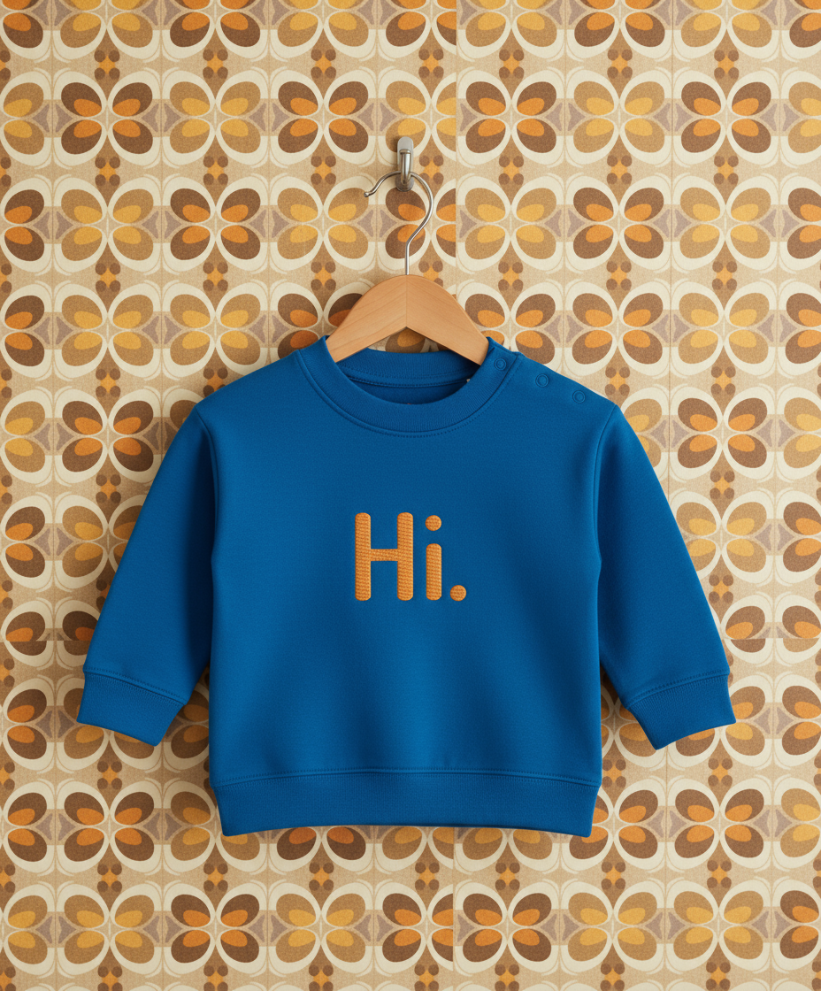 Blue sweatshirt with 'Hi.' embroidered on it hanging on a wooden hanger against a patterned wall.